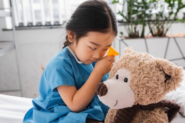 little asian girl playing in clinic and examining teddy bear with toy otoscope clipart