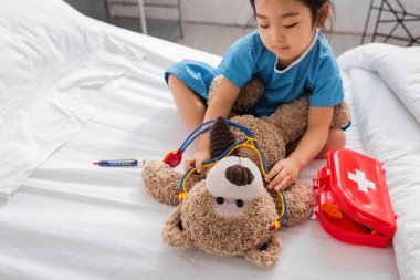 Pediyatri kliniğinde yatakta oynarken oyuncak steteskop takan Asyalı kız.