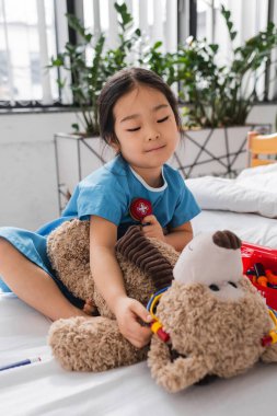 Hastane cüppeli gülümseyen Asyalı kız pediatri kliniğindeki yatakta oyuncak ayı ve steteskopla oynuyor.