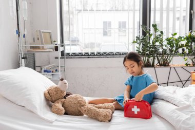 Modern pediyatri kliniğinde oyuncak ilk yardım çantasını oyuncak ayının yanında açan Asyalı kız.