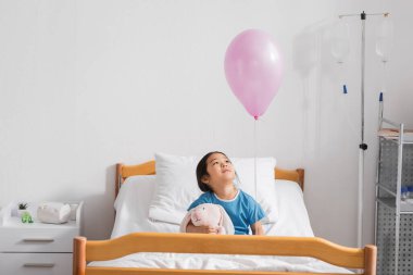 joyful asian girl sitting on hospital bed with toy bunny and looking at festive balloon clipart