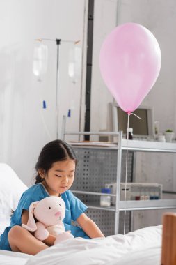 Küçük Asyalı çocuk hastane yatağında balonun yanında oyuncak tavşanla oynuyordu.