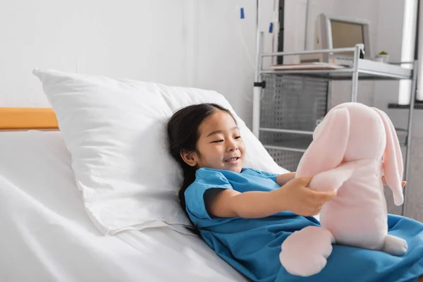 穿着医院长袍的快乐的亚洲女孩躺在病床上和玩具兔子玩耍 — 图库照片