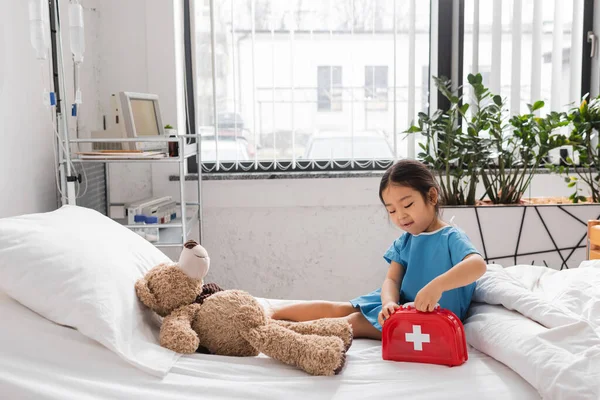 asian girl opening toy first aid kit near teddy bear on bed in modern pediatric clinic