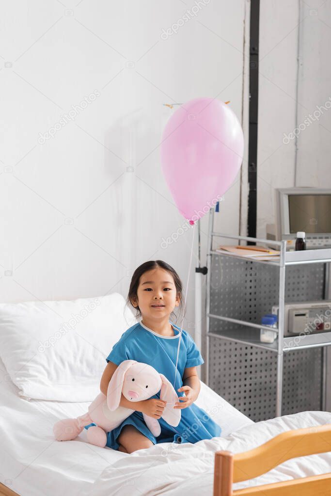 Carefree asian girl smiling at camera while sitting with balloon and toy bunny on hospital bed