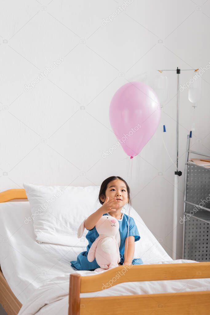 Asian girl sitting with toy bunny on hospital bed and looking at balloon