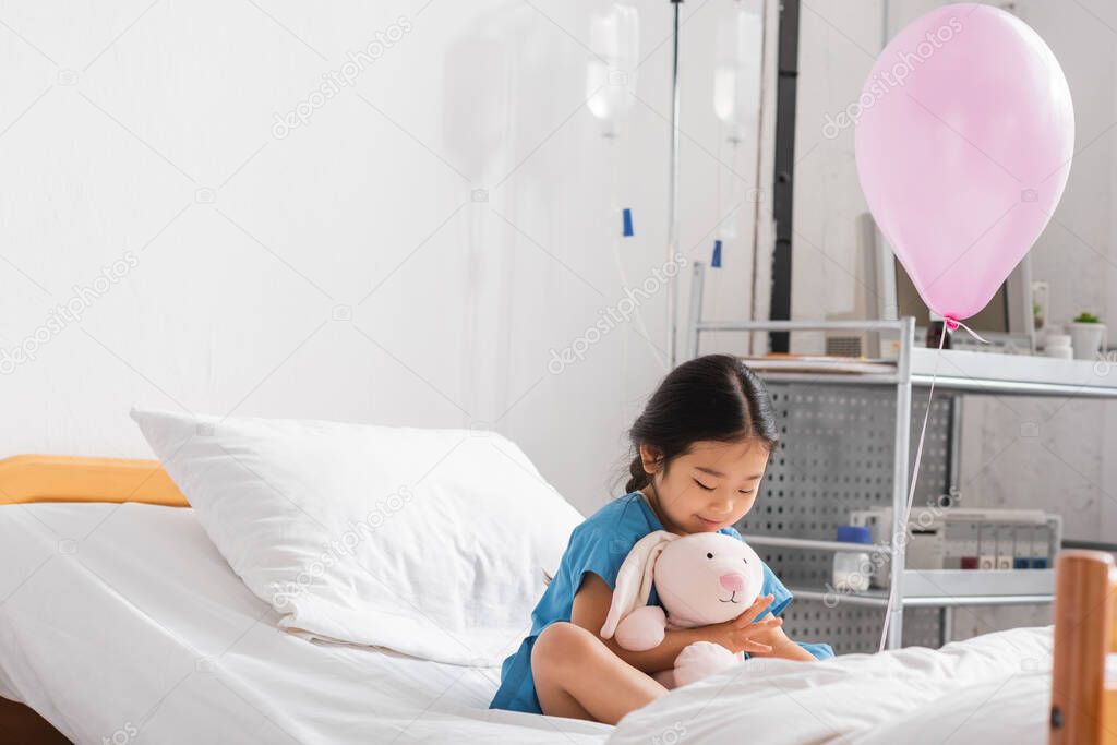 Cheerful asian girl playing with toy bunny near festive balloon on hospital bed
