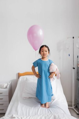 full length of asian girl in hospital gown standing with toy bunny and festive balloon on bed in pediatric clinic clipart