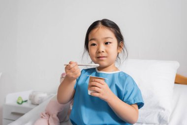 positive asian child in hospital gown eating delicious yogurt in clinic clipart