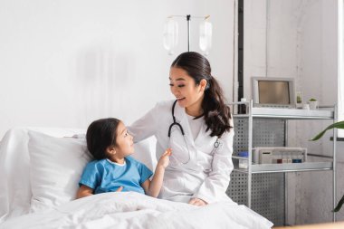 Steteskoplu mutlu Asyalı doktor hastane koğuşundaki kızla konuşuyor.