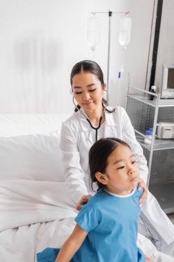 Steteskoplu gülümseyen Asyalı doktor hastanedeki küçük hastayı muayene ediyor.