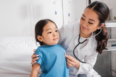 Pediatri kliniğinde steteskoplu küçük Asyalı kızı muayene eden neşeli bir doktor.