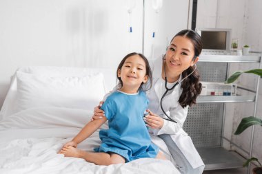 Steteskoplu gülümseyen Asyalı doktor pediatri kliniğindeki küçük kızı incelerken kameraya bakıyor.