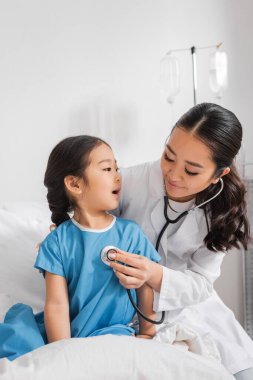 Hastane koğuşunda steteskoplu gülümseyen bir çocuk doktoruyla konuşan Asyalı bir kız.