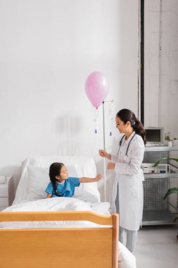 Beyaz önlüklü gülümseyen doktor hastane yatağında Asyalı bir kızın yanında balonla duruyor.