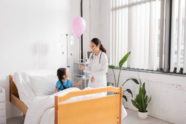Gülümseyen Asyalı doktor, modern hastane koğuşunda yatağın yanında balon tutuyor.