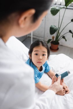 Hasta koğuşundaki şaşırmış Asyalı çocuğa astım ilacı gösteren bulanık bir çocuk doktoru.