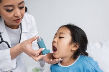Genç Asyalı doktor pediatri kliniğinde ağzı açık küçük bir kızın yanında astım spreyi tutuyor.