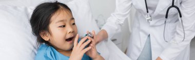 Asyalı çocuk astım spreyi tutuyor ve hastane koğuşunda beyaz önlüklü doktorun yanında ağzını açıyor.
