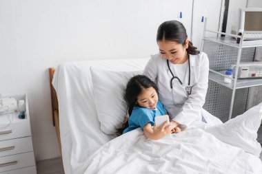 Hastane koğuşunda gülümseyen Asyalı bir doktorla akıllı telefondan selfie çeken kaygısız bir çocuk.