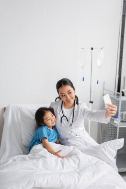 Beyaz önlüklü mutlu doktor, hastane koğuşunda pozitif Asyalı çocukla selfie çekiyor.