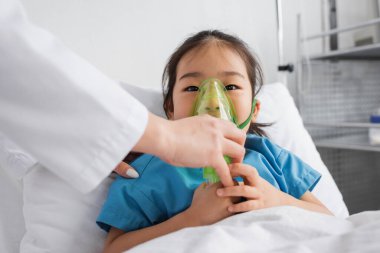 little asian girl breathing in oxygen mask near doctor in hospital ward clipart