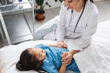 Pediyatri kliniğinde küçük bir hastanın elini tutan beyaz önlüklü Asyalı doktor.