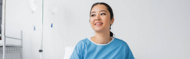 Hastane önlüklü genç Asyalı kadın klinikte gülümsüyor ve yüzünü çeviriyor.
