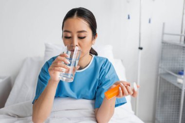 Genç Asyalı kadın klinikteki yatağın üzerinde ilaç kutusunu tutarken su içiyor.
