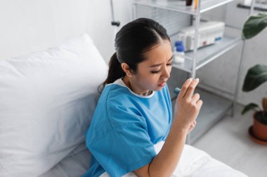 Hasta Asyalı kadının hastane koğuşunda yatarken astım spreyi tutarken yüksek açılı görüntüsü.