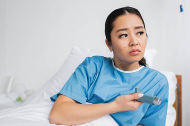 Üzgün ve düşünceli Asyalı kadın astım spreyi tutuyor ve hastaneye bakıyor.