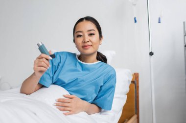 Hastane koğuşunda astım spreyini tutarken kameraya gülümseyen pozitif Asyalı kadın