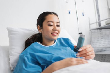 Hastane önlüklü mutlu Asyalı kadın klinikte yatakta uzanıyor ve astım spreyine bakıyor. 