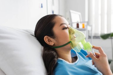 Hastane koğuşunda yatan oksijen maskeli hasta Asyalı kadının yan görüntüsü.