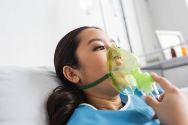Oksijen maskeli genç Asyalı kadın hastane yatağında uzanıyor ve başka tarafa bakıyor.