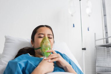 Hasta Asyalı kadın, klinikte gözleri kapalı yatarken oksijen maskesi soluyor.