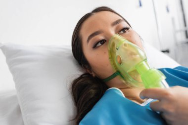 Hasta Asyalı kadın oksijen maskesi takmış yatıyor ve hastane koğuşuna bakıyor.