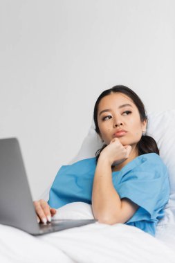 Düşünceli Asyalı kadın hastane yatağında dizüstü bilgisayarla oturuyor ve başka tarafa bakıyor.
