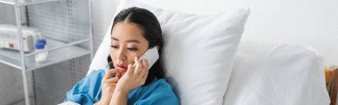 Endişeli Asyalı kadın hastane koğuşunda akıllı telefondan konuşuyor ve el ele tutuşuyor.