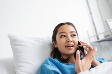 şaşırmış Asyalı kadın cep telefonuyla konuşuyor ve hastane koğuşunda el kol hareketi yapıyor.