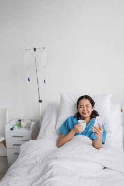 excited asian woman with smartphone and cup of tea laughing with closed eyes on hospital bed clipart