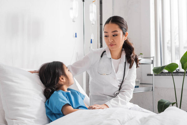 молодой азиатский педиатр со стетоскопом, смотрящий на девушку на кровати в больничном отделении