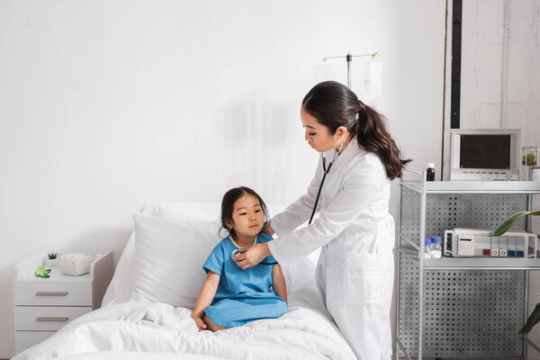 pediatrician in white coat examining asian girl with stethoscope in hospital ward