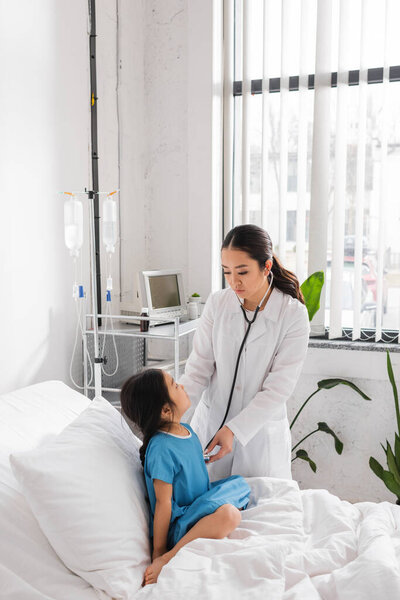 asian pediatrician with stethoscope examining little patient sitting on hospital bed