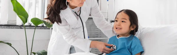 doctor with stethoscope examining cheerful asian girl in pediatric hospital, banner