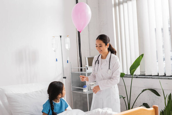 cheerful asian pediatrician holding festive balloon near little patient sitting on bed in clinic