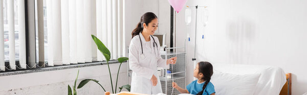 happy asian doctor in white coat holding festive balloon near kid in modern pediatric clinic, banner