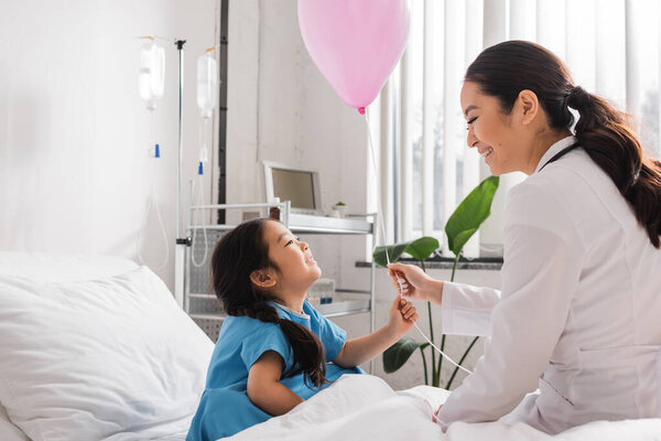 side view of young pediatrician giving festive balloon to overjoyed asian girl in hospital ward
