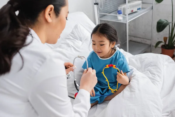 Размытый Доктор Показывает Стетоскоп Положительной Азиатской Девушке Сидящей Игрушкой Больничной — стоковое фото