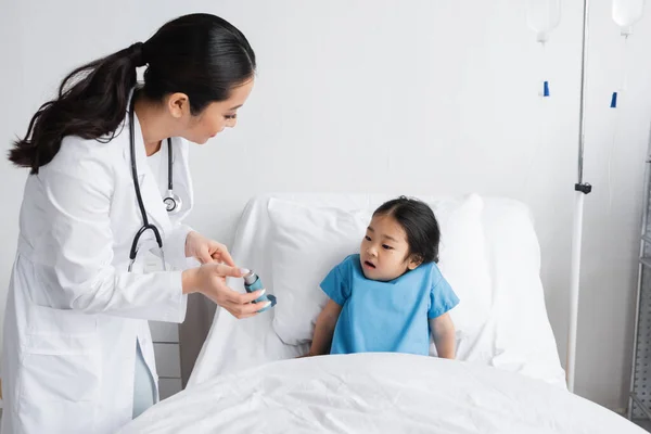 Ung Läkare Visar Inhalator Till Förvånad Asiatisk Flicka Sitter Sängen — Stockfoto